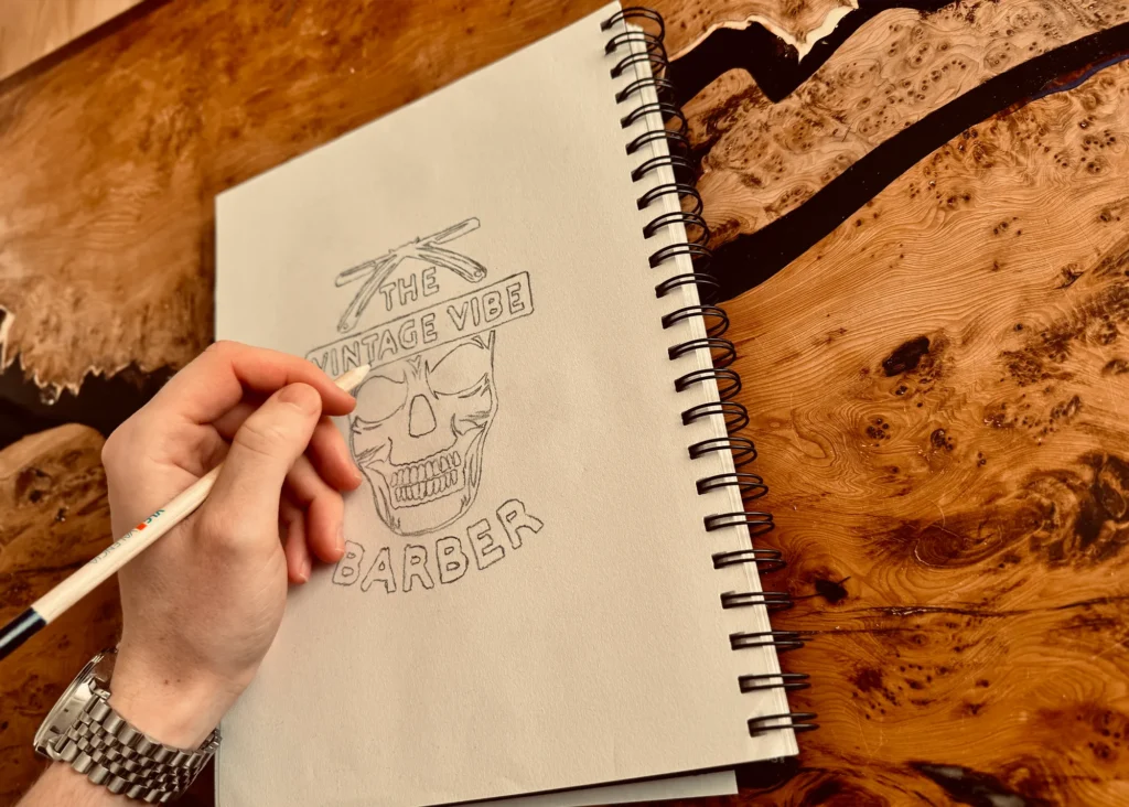 Hand drawing a vintage-inspired barber logo featuring a skull and crossed razors in a sketchbook on a rustic wood table.