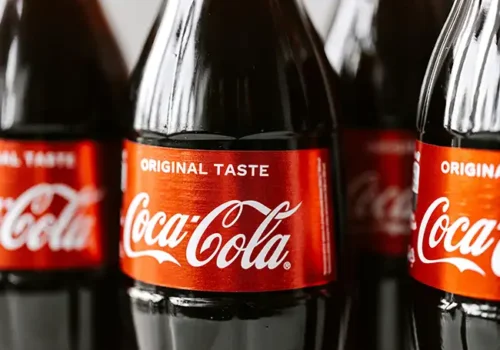 Close-up of several Coca-Cola glass bottles with red labels displaying the iconic 'Coca-Cola' logo and 'Original Taste' text, emphasising the brand's classic beverage.
