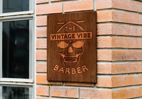 Wooden sign for 'The Vintage Vibe Barber' featuring a carved skull with sunglasses and crossed straight razors, mounted on a brick wall outside a barber shop.