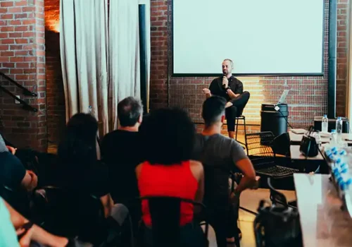 Speaker sitting on a stool and addressing an audience in a brick-walled venue, with attendees seated and listening attentively, creating an intimate and engaging presentation environment.