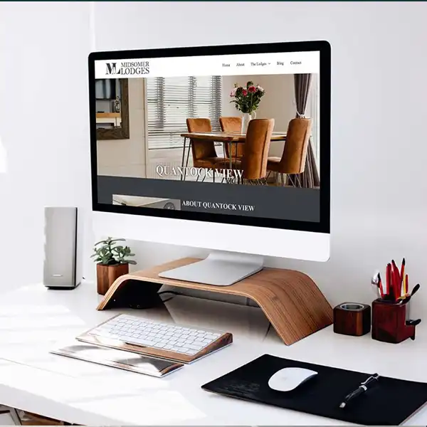 Modern home office setup with a desktop computer displaying a website featuring a luxury lodge. The workspace includes a sleek white desk with stylish accessories like a wood monitor stand, a small plant, and office supplies.