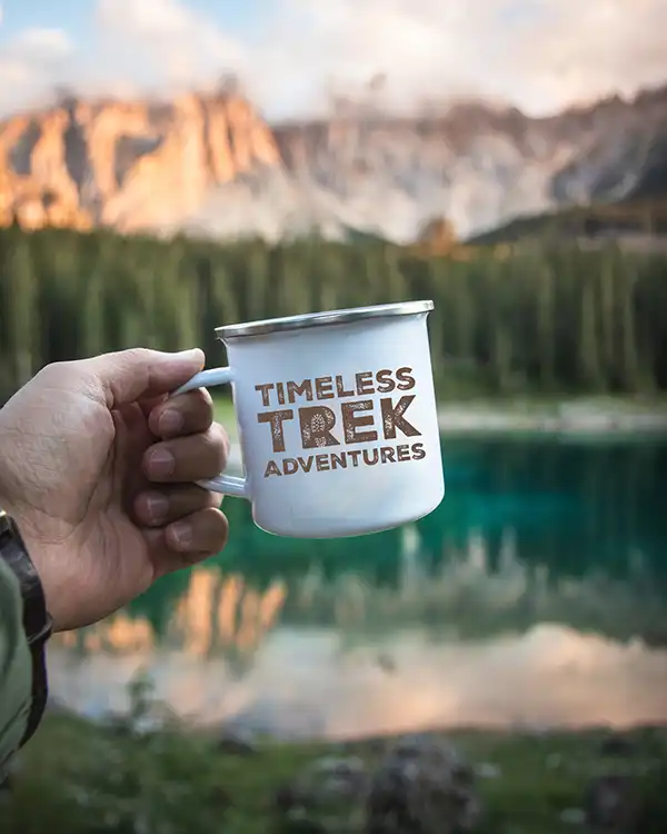 Hand holding a white enamel mug with 'Timeless Trek Adventures' printed in a rustic font, set against a scenic backdrop of a mountain lake and forest at sunset.